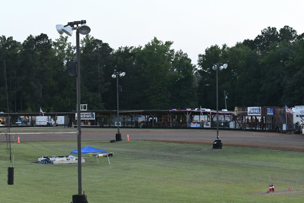 Outdoor dirt racetrack with light poles, grassy infield, a small blue tent, and a covered structure with signs and people in the background, surrounded by trees.