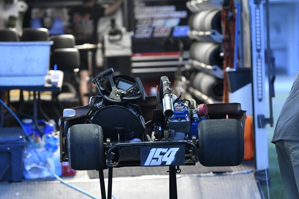A go-kart with number 154 is parked inside a trailer, surrounded by tires and equipment, with a mechanic partially visible on the right.