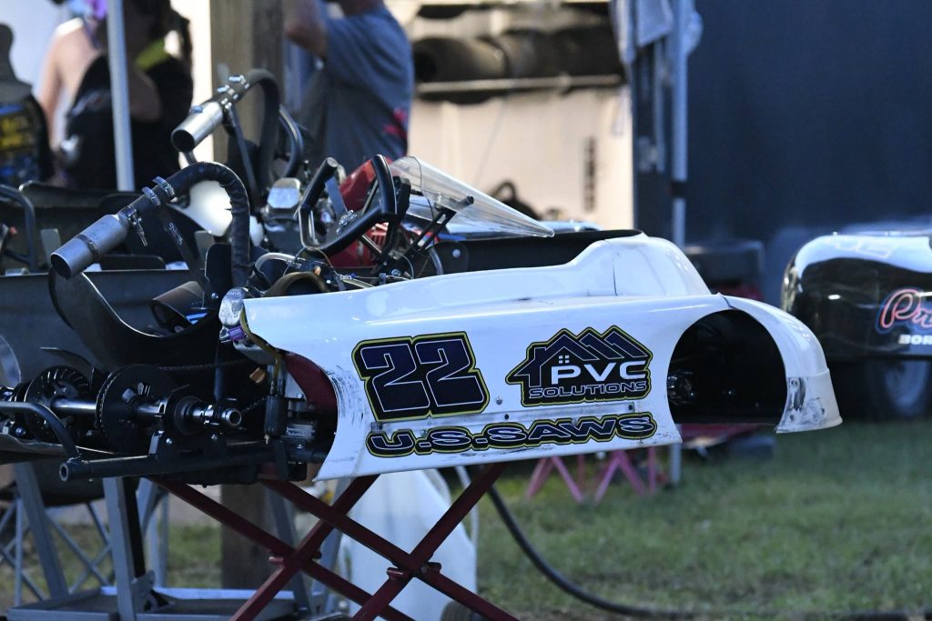 Go-kart with number 22 and PVC Solutions sponsor logo, parked on a stand outdoors with mechanic equipment and people in the background.