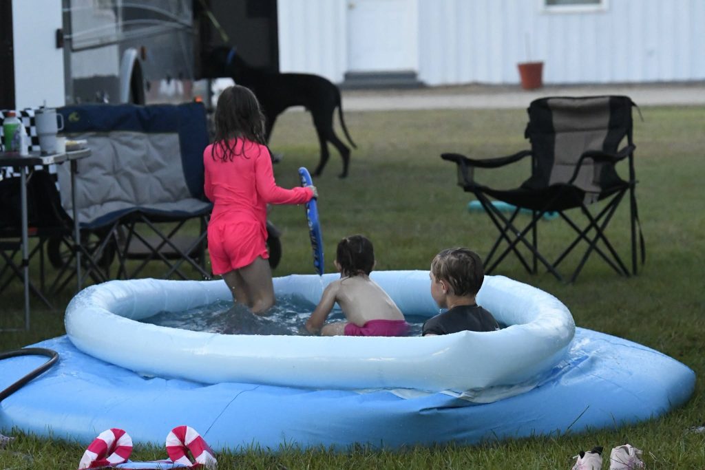 Three children playing in an inflatable pool outdoors, with lawn chairs and a black dog nearby, suggesting a casual summer day.