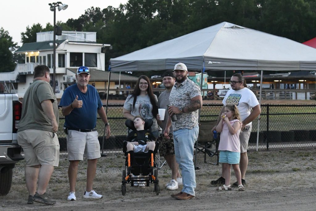 A group of people, including a child in a wheelchair, stand together outdoors near a tent and building, smiling and posing for a photo.
