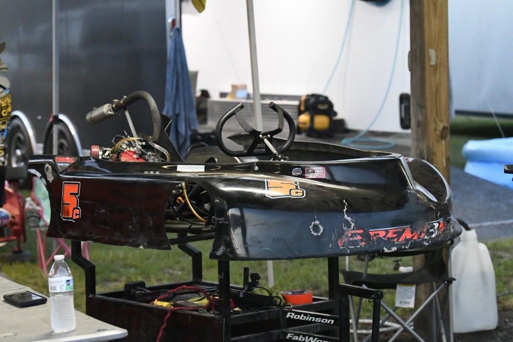 A worn black go-kart with “5C” and “Premier” labels sits on a stand in a garage area, surrounded by tools and equipment.