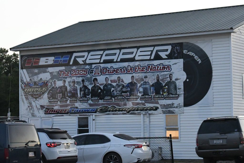 Large banner on white building displays 2025 HBM Reaper racing drivers, featuring portraits and names, with parked cars visible in front.