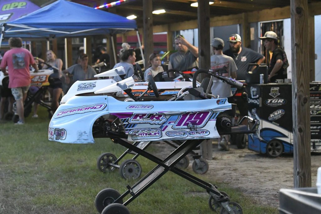 Go-karts on stands in a paddock area with people gathered under a canopy and nearby, preparing for a race event.