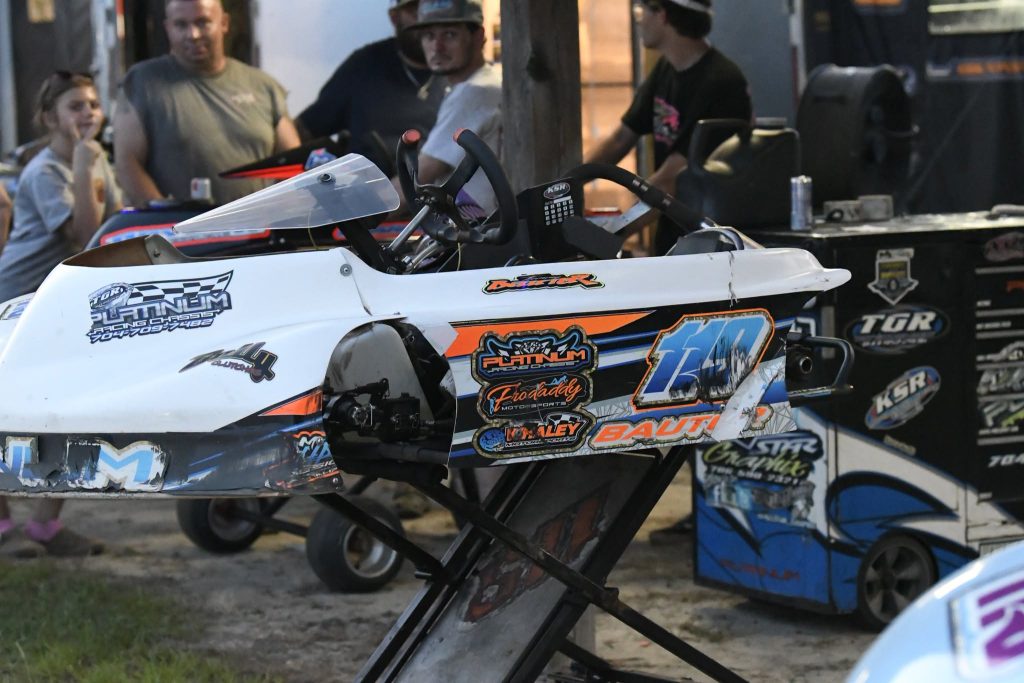 Close-up of a racing go-kart with colorful decals, surrounded by people in a pit area, tools and equipment visible in the background.