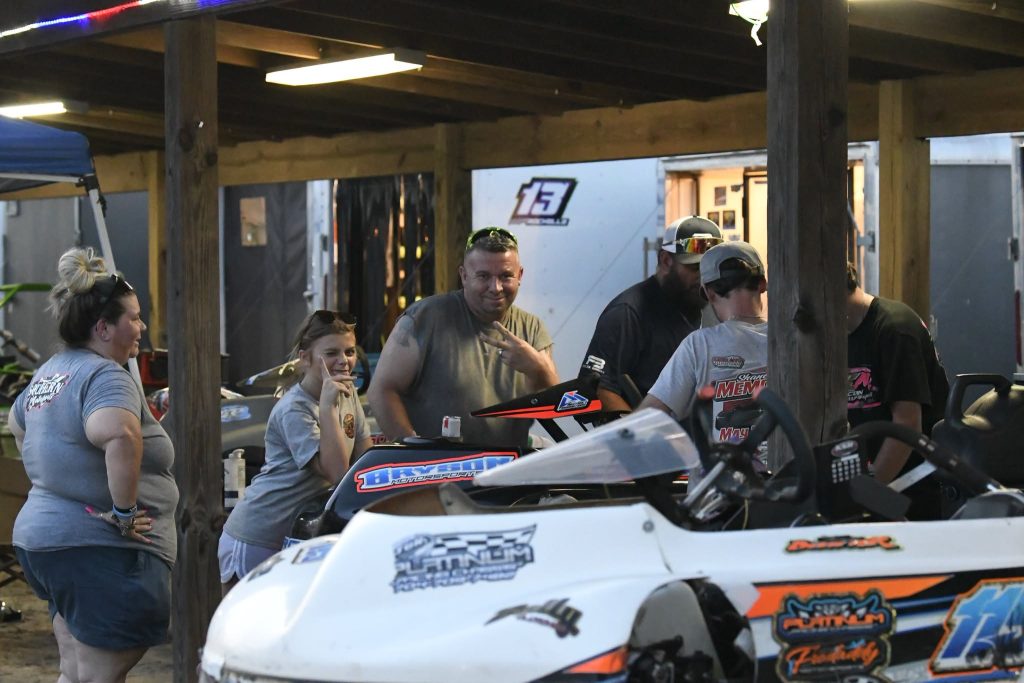 A group of people gather under a wooden shelter next to a racing vehicle, smiling and talking, with equipment and trailers in the background.