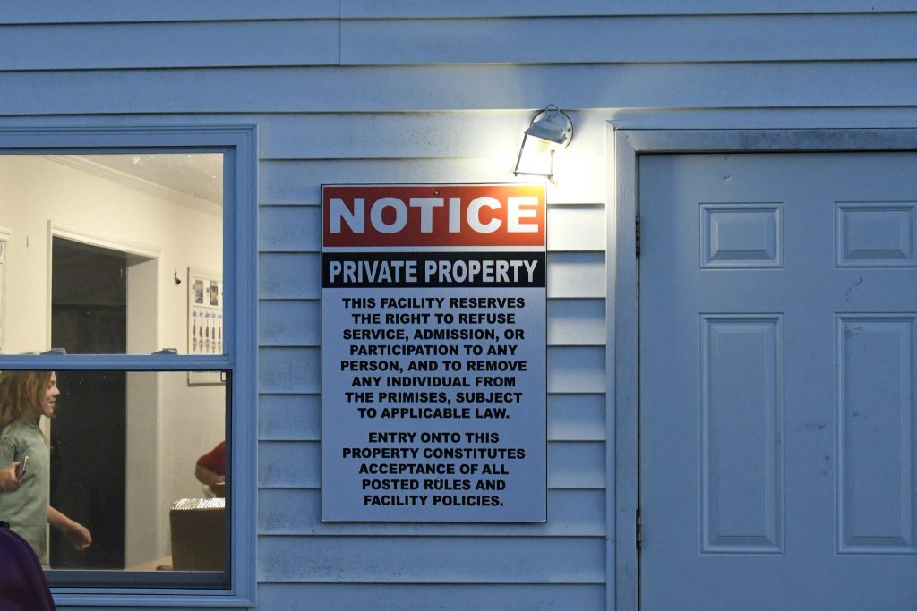 A notice sign on a building wall stating private property rules and right to refuse service, with a light above and people visible through a window.