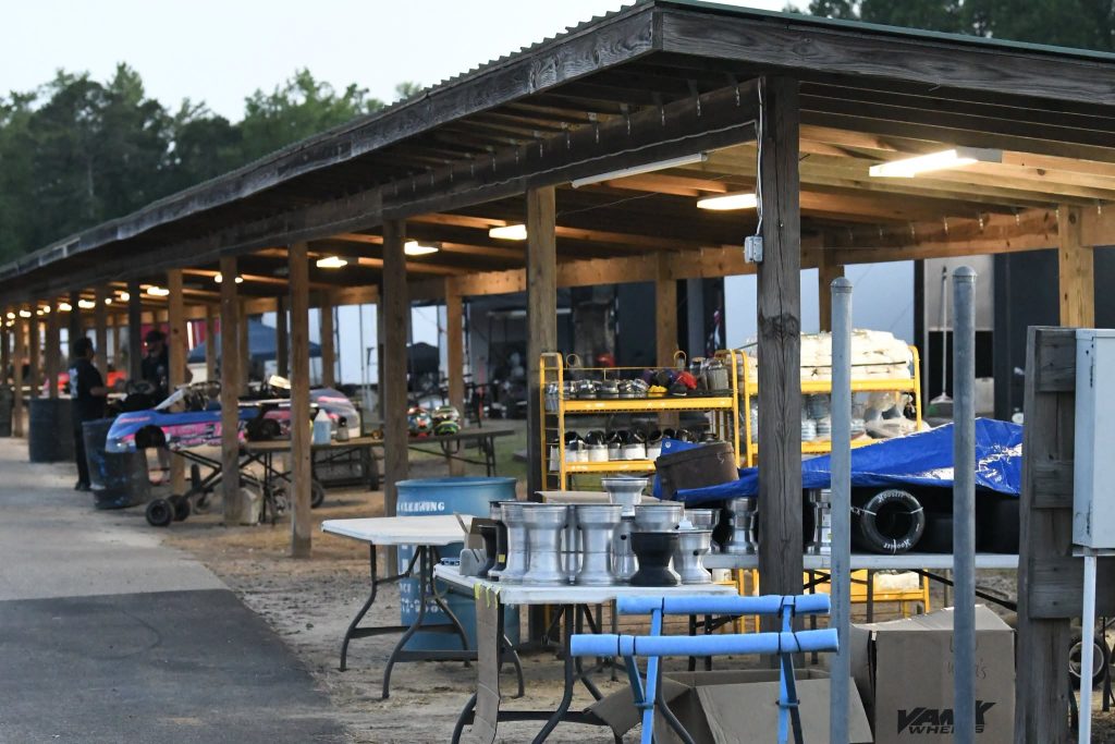Covered outdoor pit area with tools, racing kart parts, and wheels on tables and shelves, with some people and karts visible in the background.