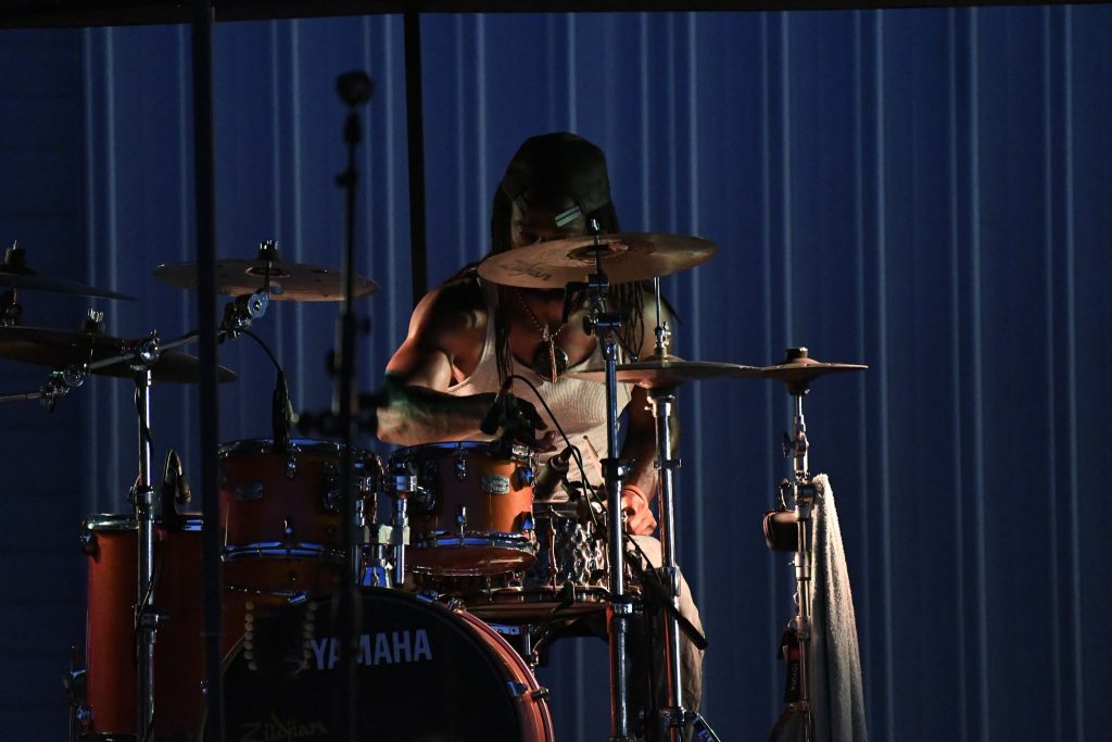 A drummer wearing a tank top and cap plays a Yamaha drum set on a dimly lit stage.