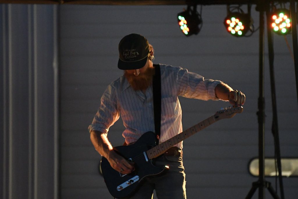 A man wearing a hat tunes an electric guitar on stage under colorful lights, partially illuminated, with a striped shirt and a dark background.