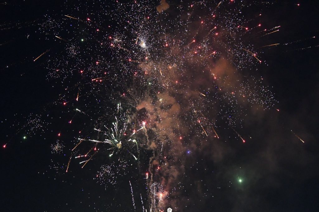 Colorful fireworks explode against a dark night sky, creating bright bursts and sparkling trails with smoke drifting through the air.
