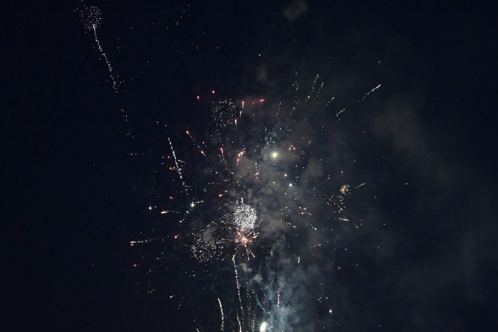 Colorful fireworks exploding in the dark night sky, with bright sparks, smoke, and scattered points of light visible.