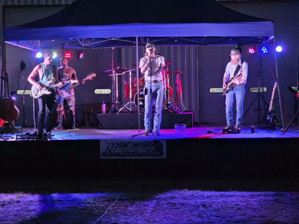 A band performs live on an outdoor stage at night, with colorful stage lighting and various musical instruments, including guitars, drums, and microphones.