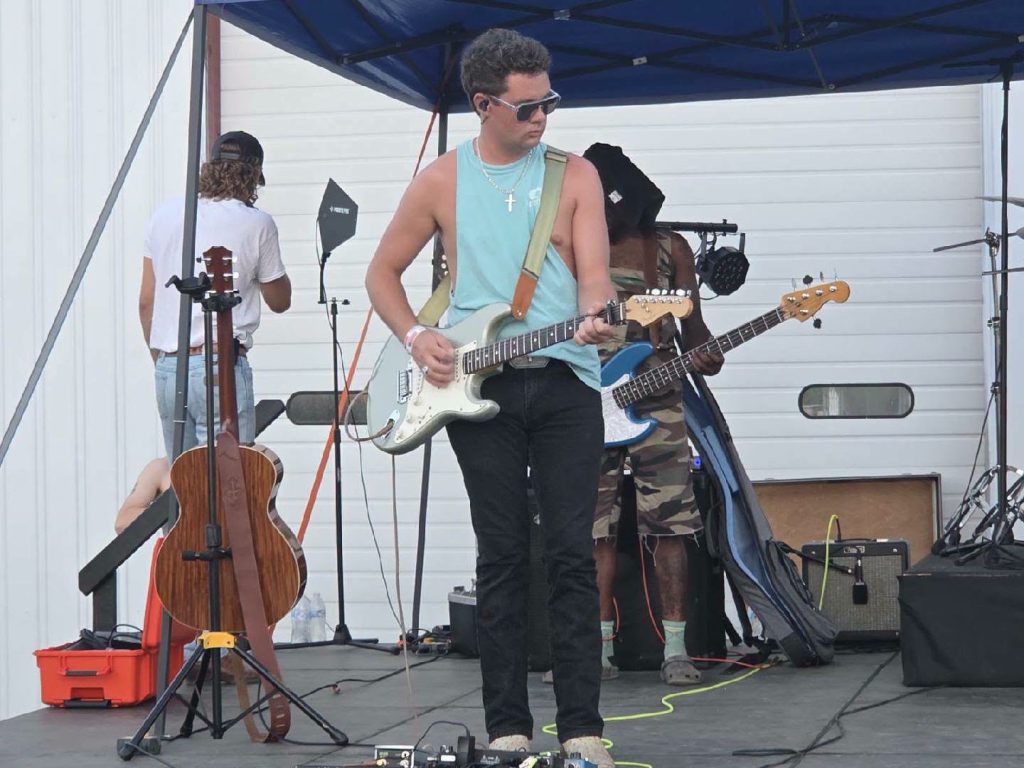 A band performs on an outdoor stage; the lead guitarist wears sunglasses and a sleeveless shirt, with other musicians and instruments visible in the background.