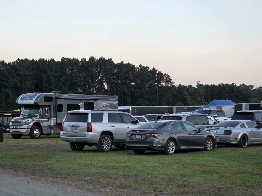 Several cars, an RV, and trailers parked on grass with trees in the background under a clear sky.