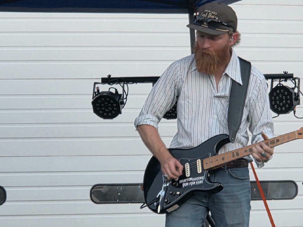 Man with a beard playing electric guitar outdoors, wearing a striped shirt, cap, and sunglasses, standing in front of a white garage door with stage lights behind him.