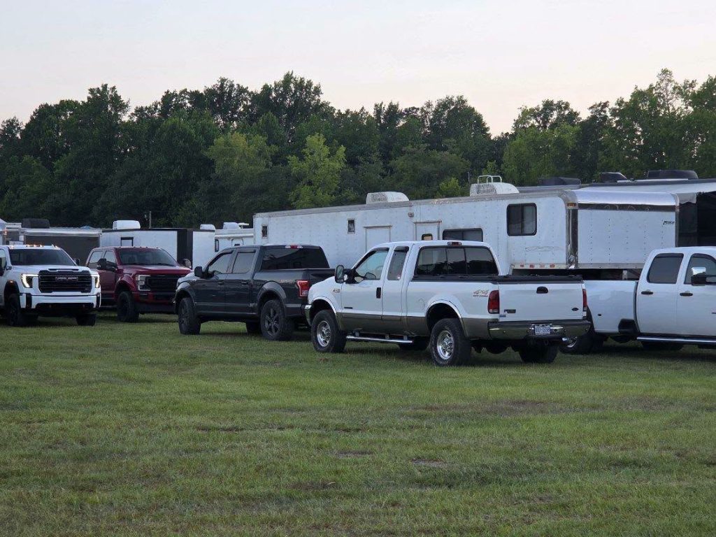 Several pickup trucks parked on grassy field beside large trailers, with trees and overcast sky in the background.