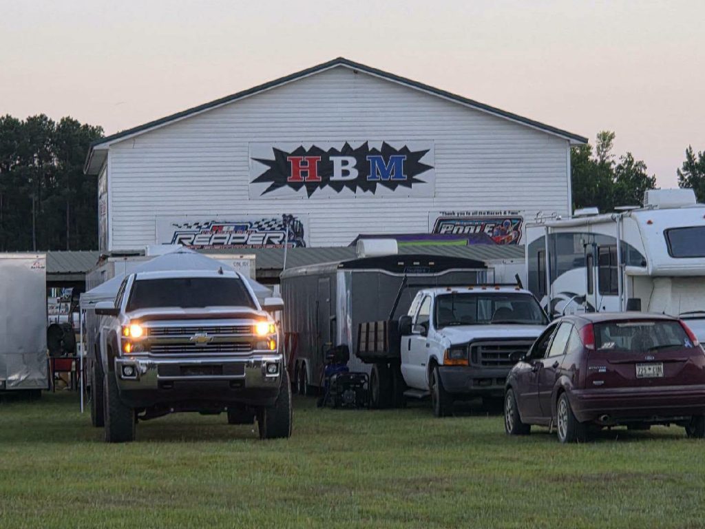 Several vehicles, trailers, and RVs are parked on grass in front of a white building with a large "HBM" sign and racing graphics.