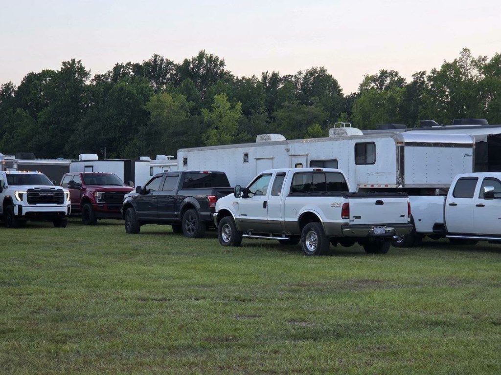 Pickups and trucks parked on grassy field in front of large white trailers, with trees and clear sky in the background.