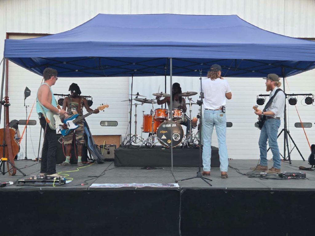 Five musicians perform on an outdoor stage under a blue canopy, featuring electric guitars, a bass, and a drum set, with amplifiers and stage equipment visible.