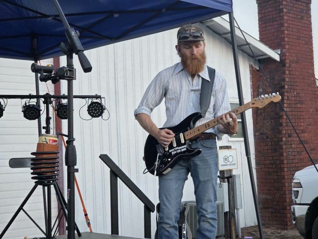 A man with a beard playing an electric guitar on an outdoor stage under a blue canopy, with stage lights and a white building in the background.
