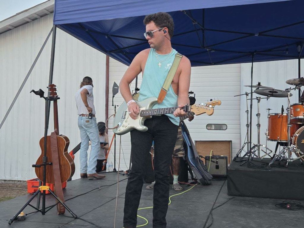 Man wearing sunglasses and sleeveless shirt plays electric guitar on outdoor stage with drums, acoustics, and equipment in background.