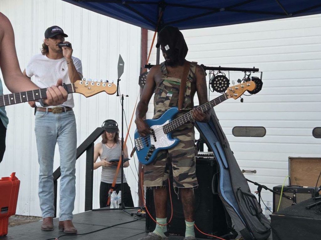 Outdoor band performance with guitarist in camouflage outfit, another musician playing guitar, a singer with a microphone, and crew member in background near equipment cases and stage setup.