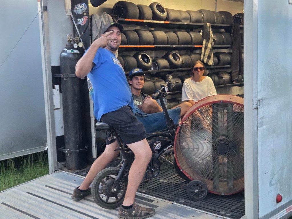 Three people relaxing inside a trailer with racing tires, a large industrial fan, and a bike. One man smiles and gestures at the camera.