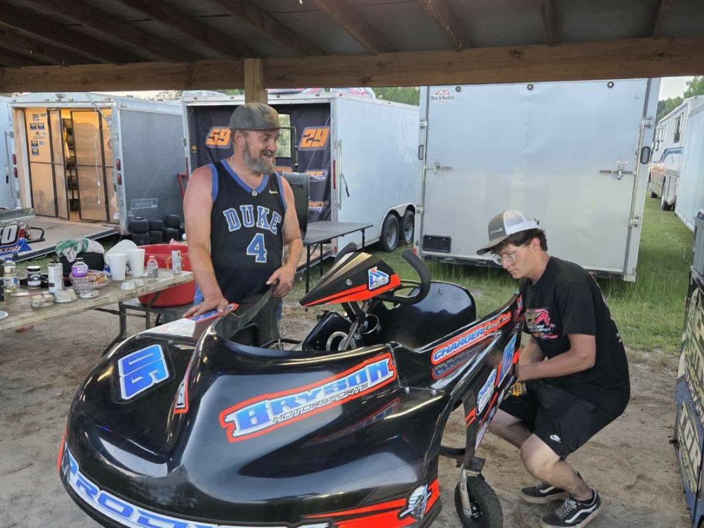 Two men work on a black racing go-kart under a canopy, with trailers and a table of tools and supplies in the background.