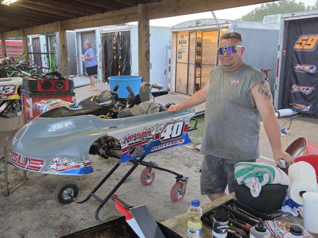 Man wearing sunglasses stands next to a racing go-kart in a pit area, surrounded by racing equipment, trailers, and tools.