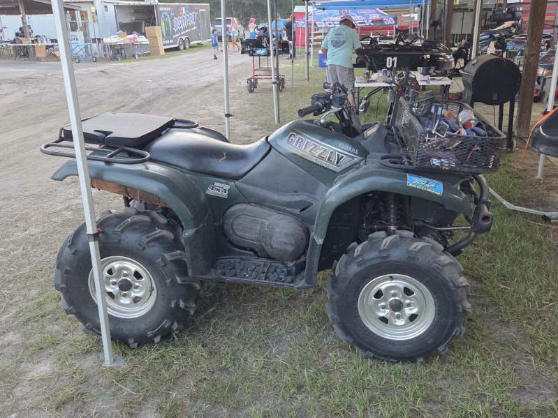 A green Yamaha Grizzly ATV parked on grass near an outdoor area with trailers and people in the background.