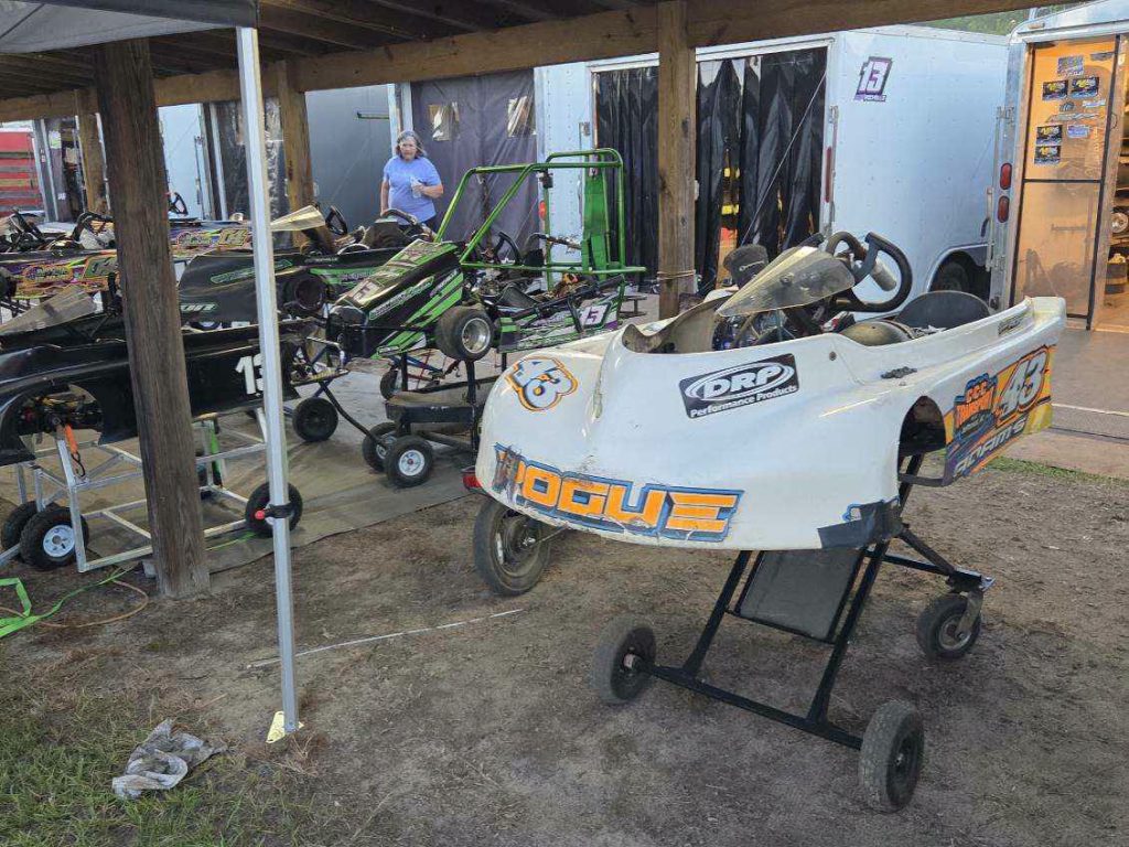 Several go-karts on stands under a wooden shelter at a race track, with trailers and a person in the background.