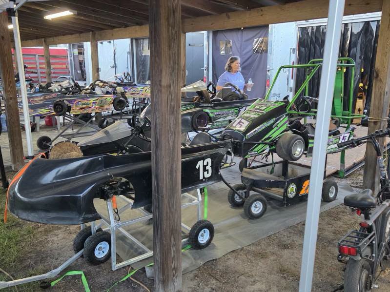 Several go-karts with numbers are parked under a wooden shelter, with a woman sitting in the background near a trailer.