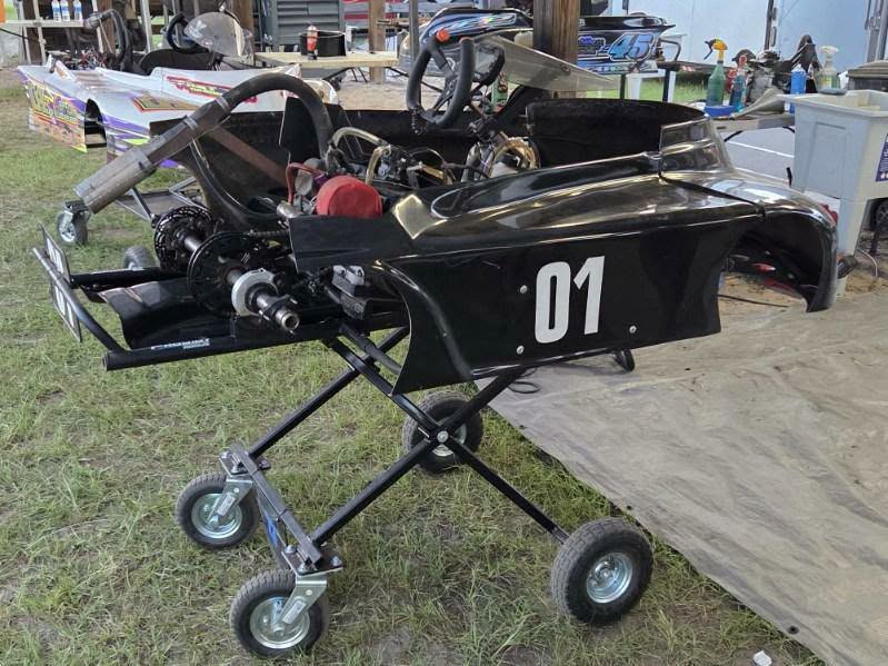 Black go-kart with number 01 displayed, elevated on a maintenance stand outdoors, surrounded by other racing karts and equipment.
