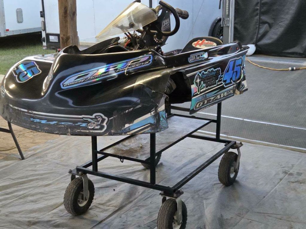 A black racing go-kart with number 45 is displayed on a wheeled metal stand inside a garage or pit area.