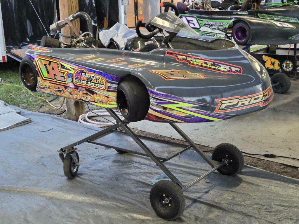 A decorated racing go-kart on a metal stand, with vibrant decals and sponsor stickers, in a pit area with other karts nearby.