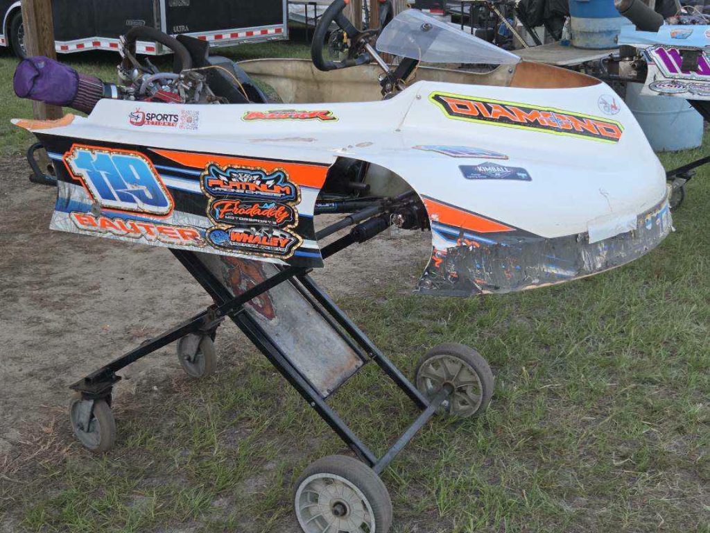 A white go-kart with racing decals and the number 119 is displayed on a wheeled stand outdoors, with grass and trailers in the background.