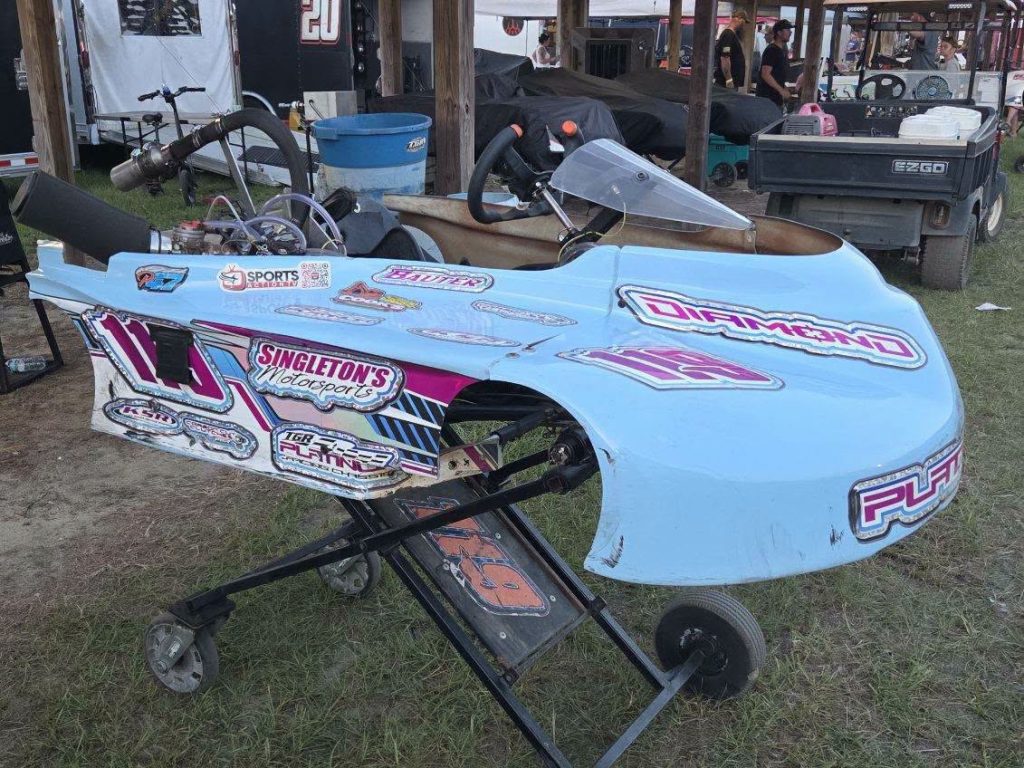 A blue go-kart with racing decals and sponsor stickers sits on a stand outdoors at a motorsports event.