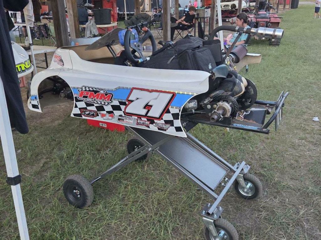 A go-kart with the number 71 is placed on a stand outdoors at a race event, surrounded by equipment and people in the background.