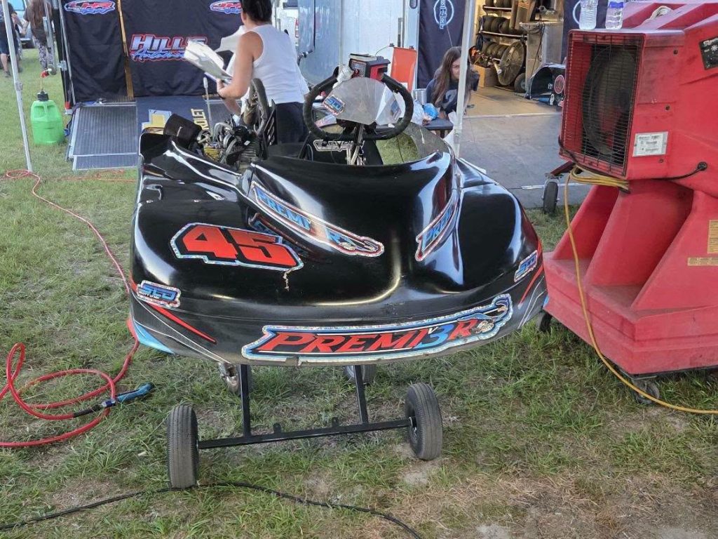 A black go-kart with "45" and "PREMI3R" decals is parked on a stand in a pit area, surrounded by equipment and people.