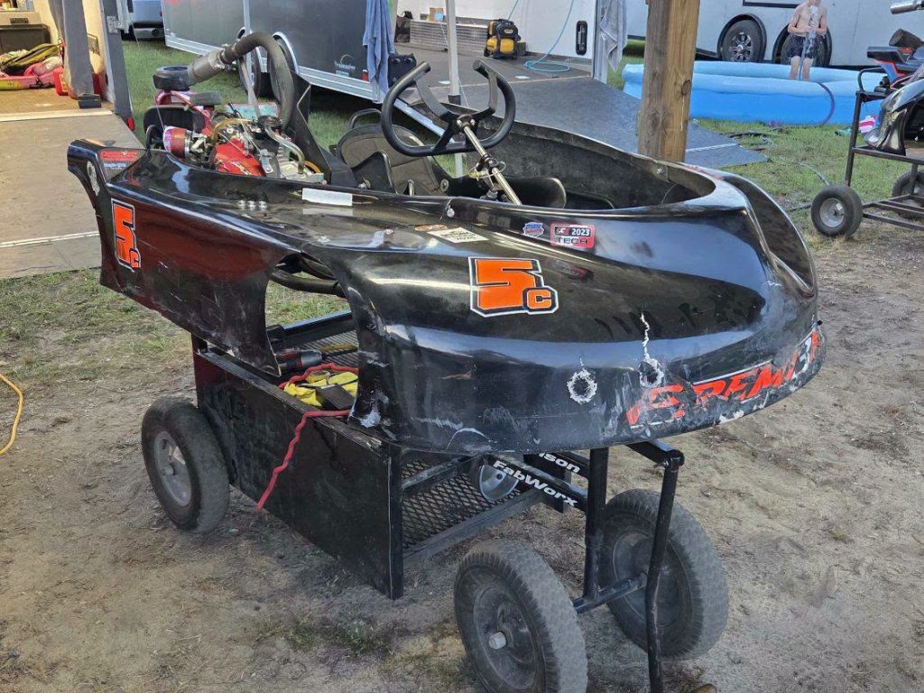 A black go-kart with visible wear and number 5C is placed on a cart in an outdoor area, with trailers and a small pool in the background.