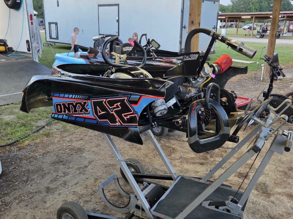 Black go-kart with “ONYX 47” decal on a stand, outdoor pits, children playing and trailer in background, racing equipment visible nearby.