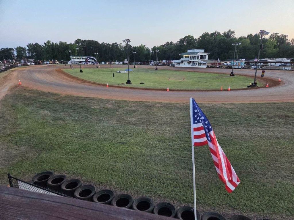 A dirt racetrack with an American flag in the foreground, surrounded by green grass, trees, and small buildings in the background.