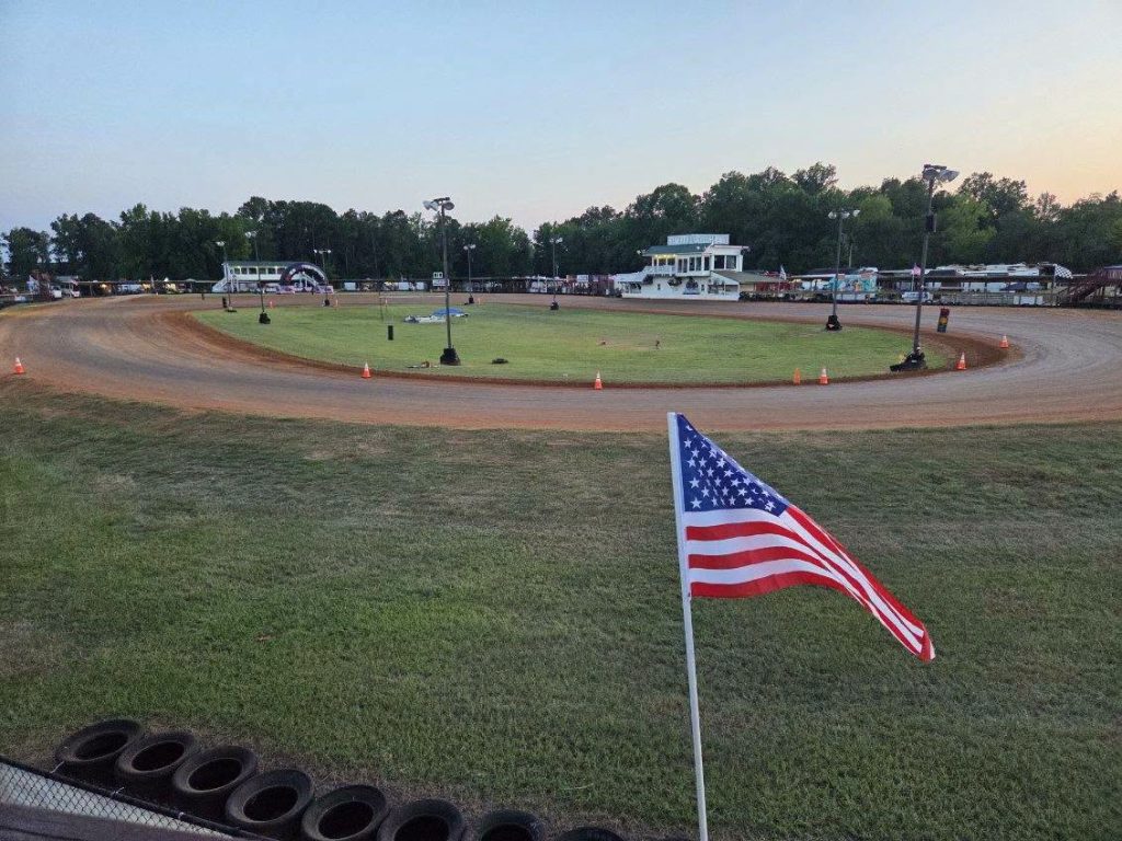 A dirt racetrack with an American flag in the foreground, surrounded by grass, light poles, and buildings, with trees and vehicles in the background.
