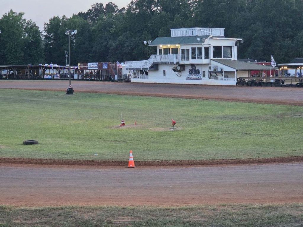 Dirt racetrack with grass infield, safety cone, tire, small flags, and a white race control building in the background, surrounded by trees.