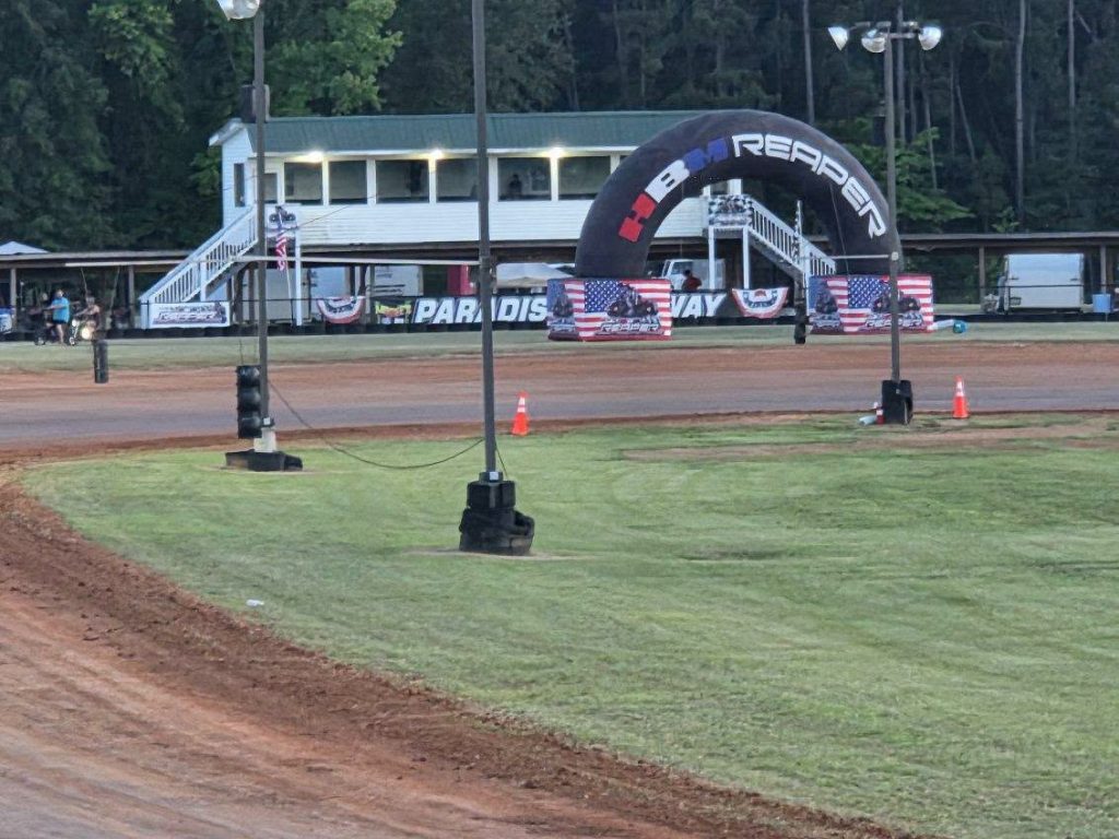 Dirt racetrack with a large arch, American flags, and a white building in the background, surrounded by green grass and some people.