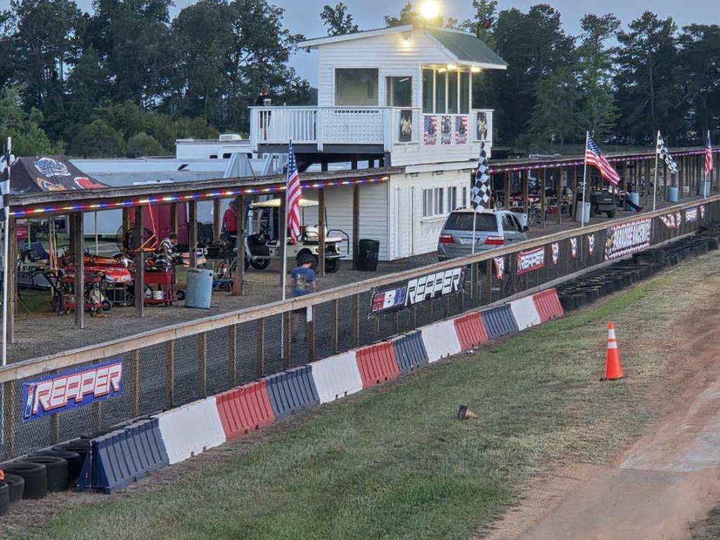 Outdoor go-kart racing track with a white control building, covered pits, American flags, checkered flags, and safety barriers along a dirt track, surrounded by trees.