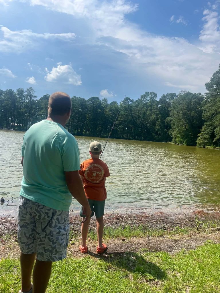 Two people, one adult and one child, standing by a lake fishing on a sunny day with trees and blue sky in the background.