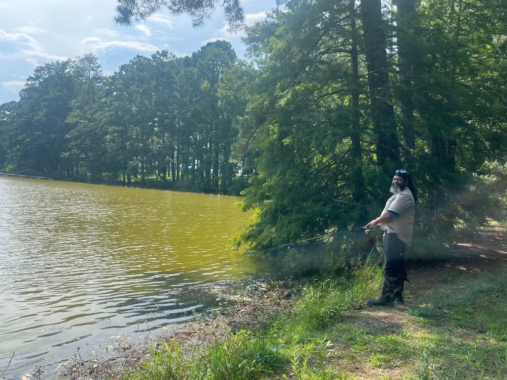 A man fishing by a lake on a sunny day, surrounded by tall green trees and calm water, with a partly cloudy sky above.