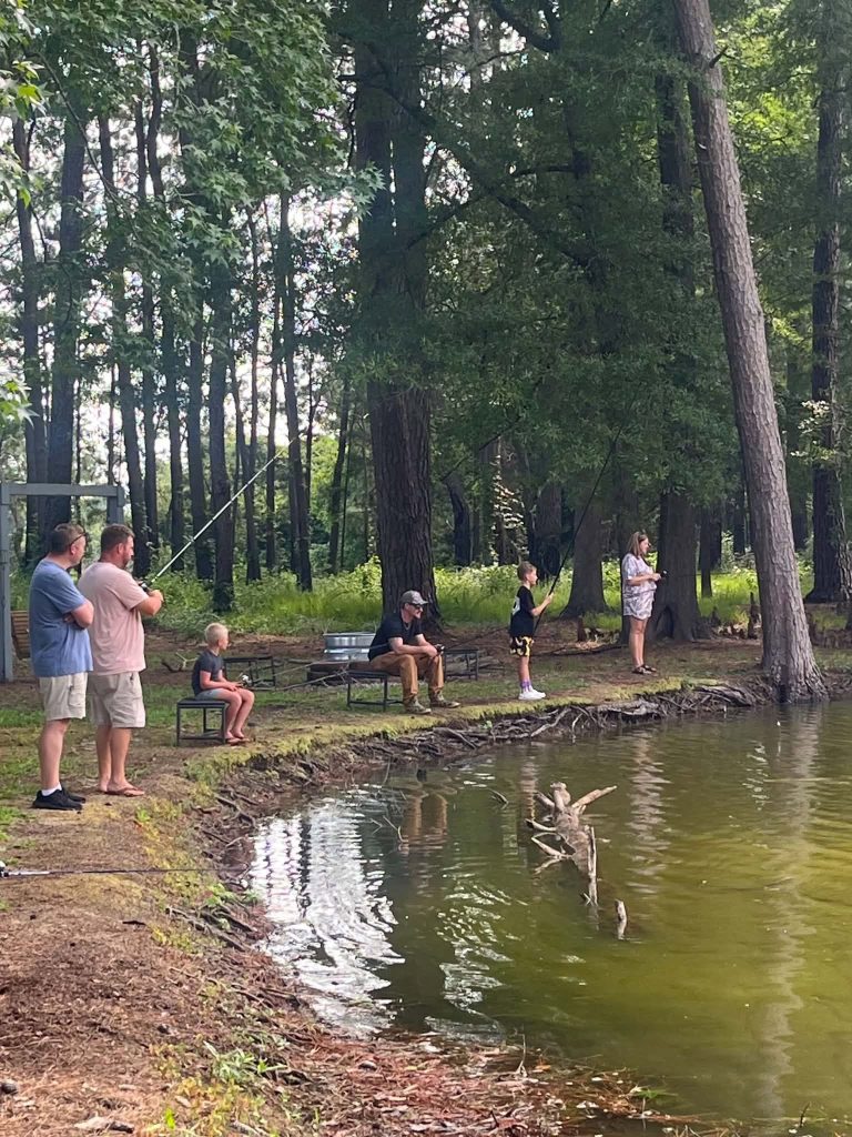 Several people, including children and adults, fishing and relaxing by a wooded pond with trees and benches along the grassy shoreline.